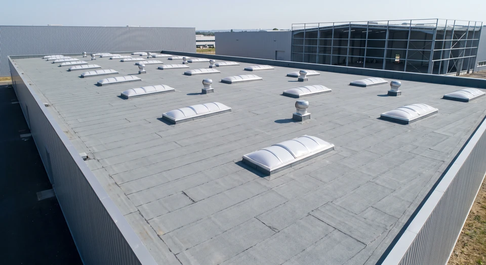 Vue aérienne d'une toiture plate industrielle large et étendue avec membrane d'étanchéité bitumineuse multicouche ou EPDM gris anthracite, montrant des joints de soudure apparents, des lanterneaux dôme en polycarbonate alignés, des relevés périmétriques, des sorties de ventilation inox et des acrotères bien définis. Structure de bâtiment industriel en arrière-plan (bac acier, bardage métallique) illustrant l'environnement professionnel et la spécialisation d'EPI en étanchéité toiture plate industrielle grande surface.