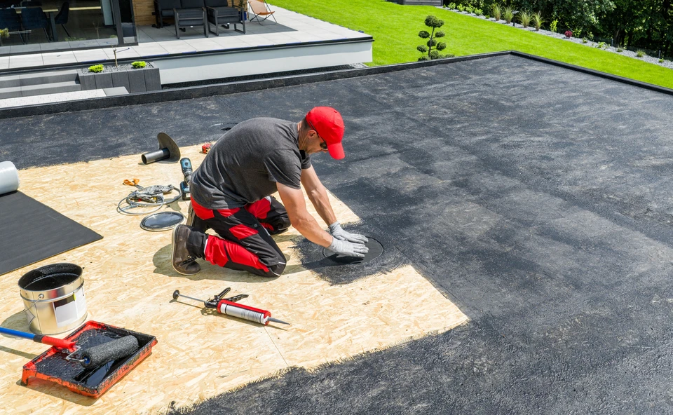 Vue en hauteur d'une toiture terrasse en cours de rénovation complète montrant plusieurs postes de travail : en premier plan, une membrane d'étanchéité bitumineuse ou PVC partiellement déroulée et en cours de pose ; au centre, des couches d'isolation thermique visibles avec chevauchement régulier des lés ; en arrière-plan, les relevés d'étanchéité périmétriques finalisés et un lanterneau dôme fraîchement installé. Structure bâtiment visible en périphérie avec acrotères en briques. La scène montre clairement l'intervention d'une seule équipe gérant plusieurs étapes techniques sur un même chantier. Lumière naturelle diurne, finition professionnelle et rigueur de pose apparentes. Pas de personnes visibles pour laisser la place au produit du travail. Aucun texte ou logo.