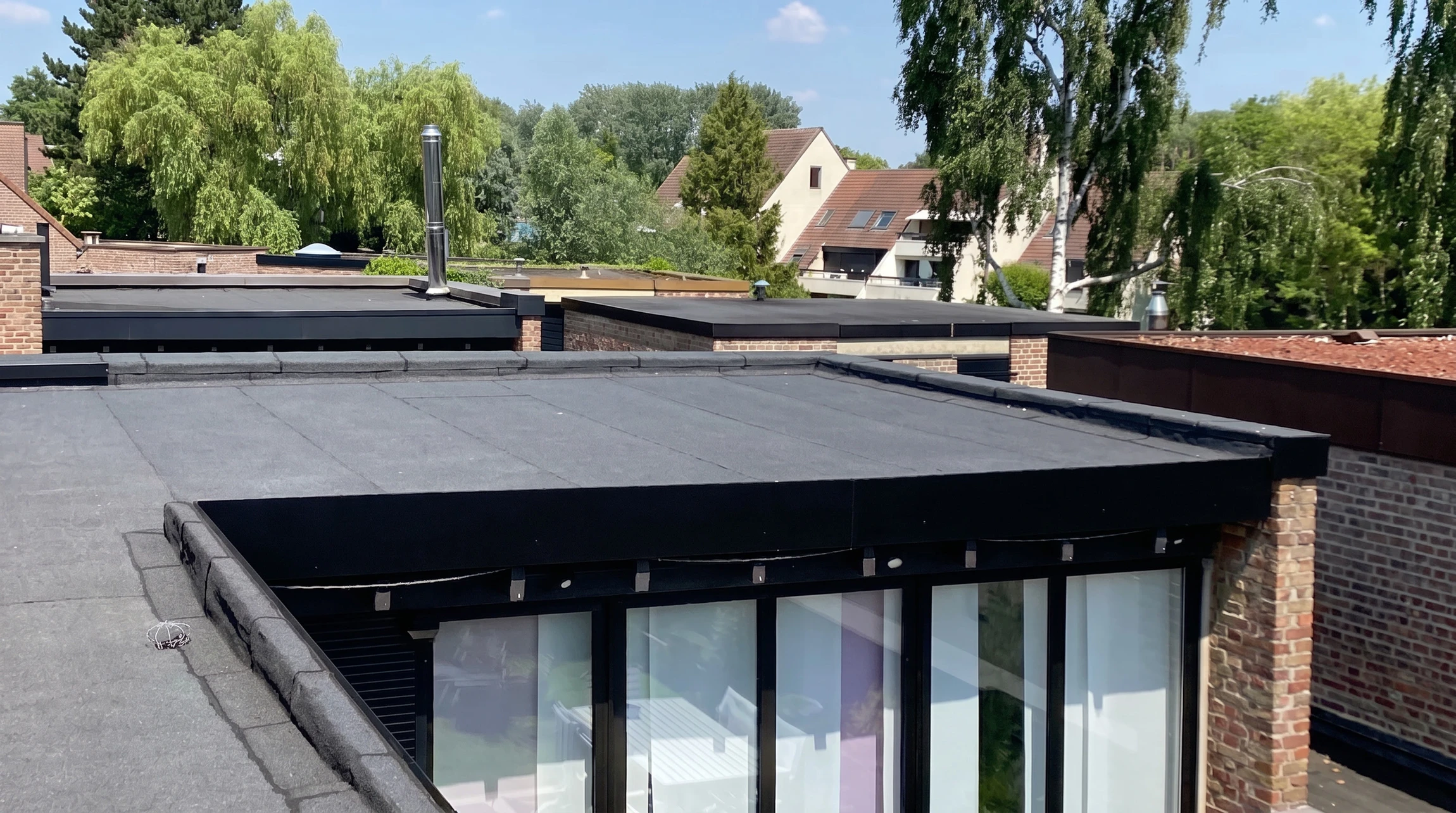 Vue aérienne d'une toiture plate en cours de réparation et d'entretien, montrant un ouvrier d'EPI effectuant un travail d'inspection ou de maintenance sur une membrane d'étanchéité bitumineuse ou EPDM bien définie. La toiture présente des relevés de membrane périphériques, des lanterneaux dôme translucides, et des finitions métalliques. L'environnement résidentiel ou petit collectif est visible en arrière-plan. L'image capture le professionnalisme et la spécialisation d'une intervention d'entretien régulier sur une toiture plate, incarnant l'expertise d'EPI.